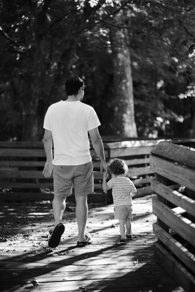Black and white image of dad walking with toddler down boardwalk